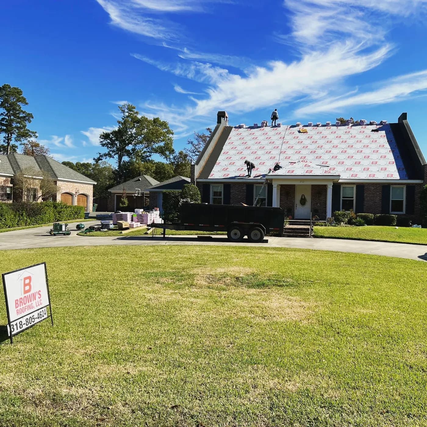 Active Brown's Roofing job site with crew on the roof, dump trailer, and a Brown's Roofing yard sign in the foreground