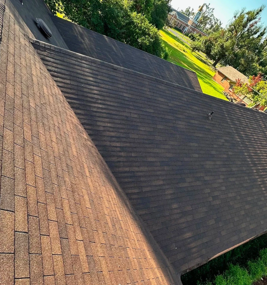 Completed brown asphalt shingle roof planes meeting at a valley on a church in Lake Providence, Louisiana, with the church grounds beyond